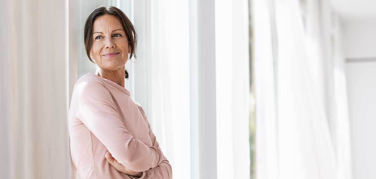 Eine Frau steht mit verschränkten Armen vor einem hohen Fenster im Wohnzimmer.